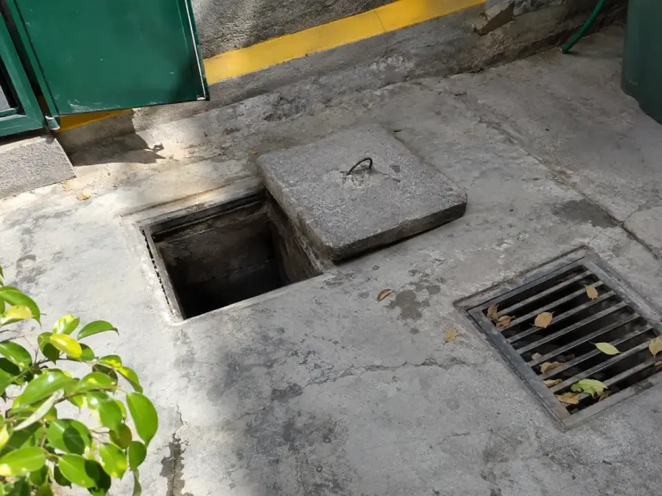 Cámara de inspección de hormigón destapada junto a la rejilla de un sumidero con hojas en un patio de servicio